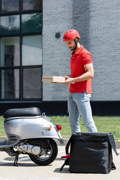 Giovane corriere musulmano in casco in possesso di scatole di pizza vicino scooter e zaino termo all'aperto — Foto stock