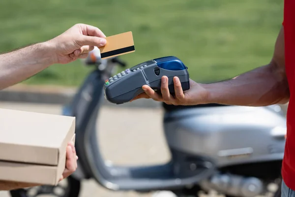 Vista recortada del hombre con tarjeta de crédito y cajas de pizza cerca de mensajero con terminal cerca de scoter borroso - foto de stock