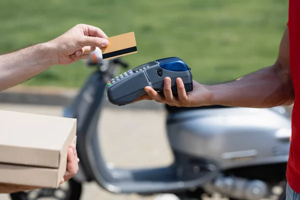 Vista ritagliata di uomo in possesso di carta di credito e scatole di pizza vicino al corriere con terminale vicino scoter sfocato — Foto stock