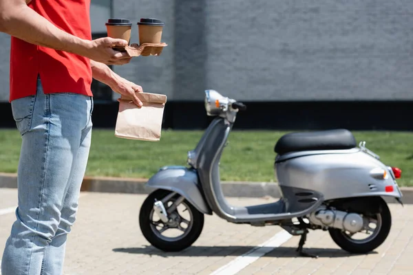 Vista recortada del mensajero en camiseta roja sosteniendo vasos de papel y bolsa cerca de scooter borroso — Stock Photo