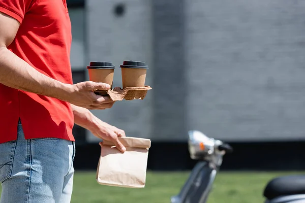 Ausgeschnittene Ansicht von Kurier mit Coffee to go und Papiertüte in der Nähe verschwommener Roller im Freien — Stockfoto