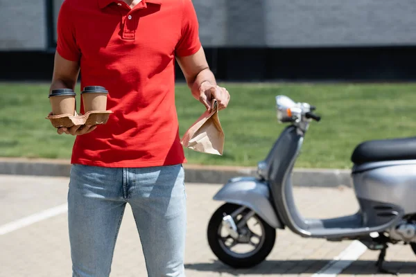 Vista ritagliata del fattorino in t-shirt rossa con sacchetto di carta e tazze vicino scooter sfocato — Foto stock