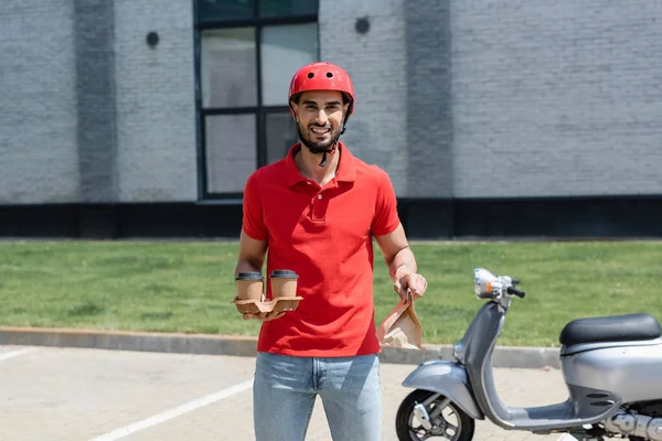 Repartidor musulmán sonriente en casco sosteniendo café para llevar y bolsa de papel cerca de scooter borroso al aire libre - foto de stock
