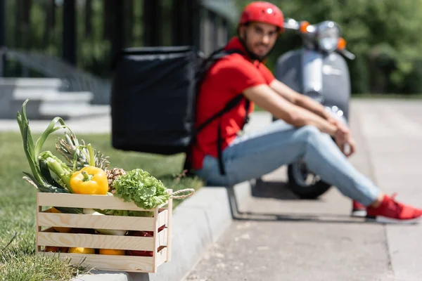 Box with fresh vegetables near blurred deliveryman and scooter outdoors — Stock Photo
