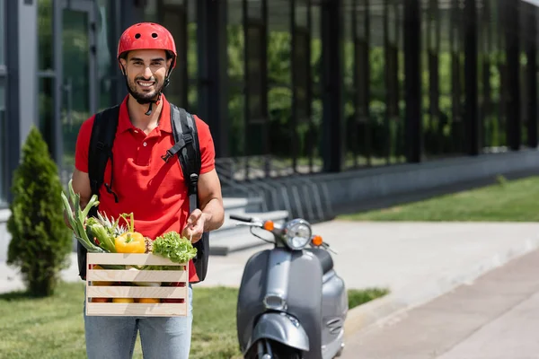 Correio árabe positivo segurando caixa de madeira com legumes frescos perto de scooter borrado — Fotografia de Stock