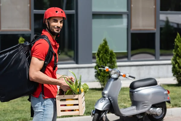 Arabischer Kurier mit Gemüse und Thermo-Rucksack lächelt in die Kamera neben verschwommenem Roller — Stockfoto