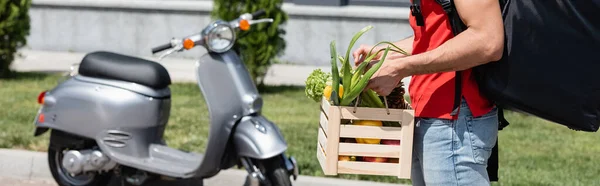 Vista ritagliata del fattorino con verdure e zaino in piedi vicino a scooter sfocato all'aperto, banner — Foto stock