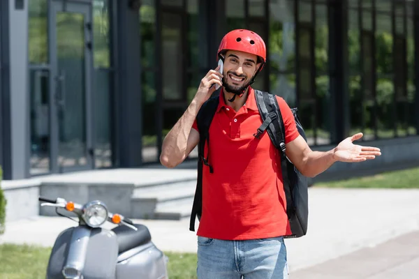 Fröhlicher muslimischer Kurier mit Thermo-Rucksack spricht auf Smartphone in der Nähe verschwommener Roller im Freien — Stockfoto