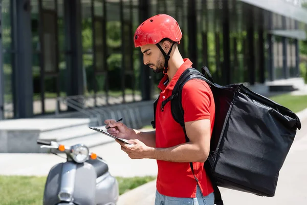 Side view of muslim deliveryman with thermo backpack holding clipboard and pen near blurred scooter outdoors — Stock Photo