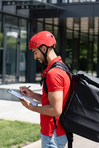 Side view of cheerful arabian courier with thermo backpack and clipboard standing outdoors — Stock Photo