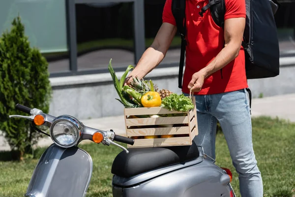Vue recadrée du livreur tenant une boîte en bois avec des légumes et de l'ananas sur scooter à l'extérieur — Photo de stock