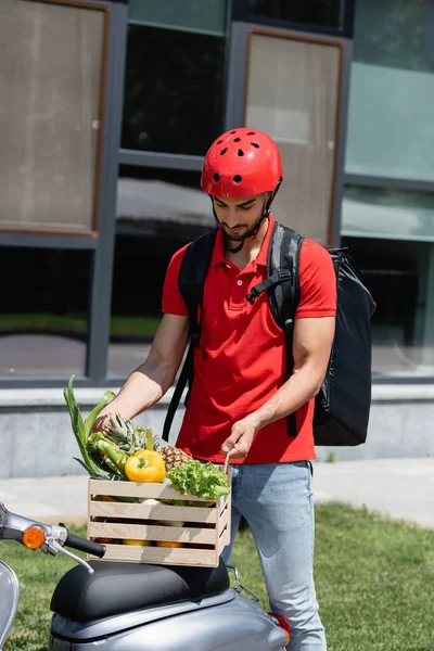 Young arabian courier in safety helmet holding wooden box with vegetables on scooter — Stock Photo