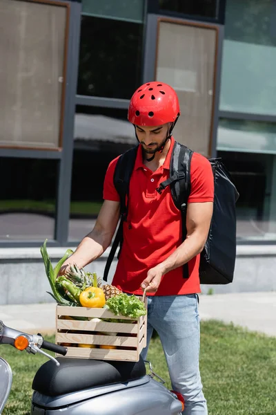 Jeune messager arabe dans un casque de sécurité tenant boîte en bois avec des légumes sur scooter — Photo de stock