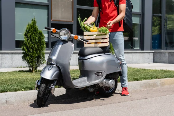 Vista cortada de correio com mochila segurando caixa com legumes orgânicos em scooter ao ar livre — Fotografia de Stock