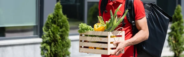 Vista recortada del repartidor sosteniendo caja de madera con verduras al aire libre, pancarta - foto de stock