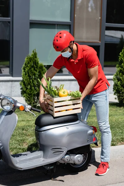 Arabe livreur dans le masque médical tenant boîte avec des légumes près de scooter à l'extérieur — Photo de stock