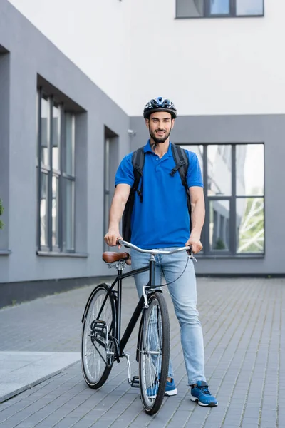 Mensajero árabe sonriente con mochila de pie cerca de la bicicleta al aire libre - foto de stock