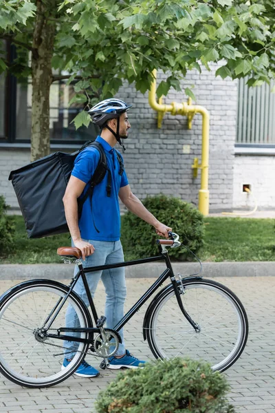 Vista lateral del mensajero árabe positivo con mochila térmica de pie cerca de la bicicleta al aire libre - foto de stock