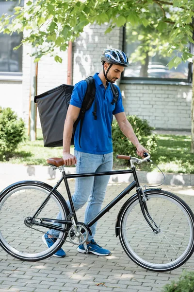 Courrier arabe avec sac à dos thermique marchant près du vélo à l'extérieur — Photo de stock