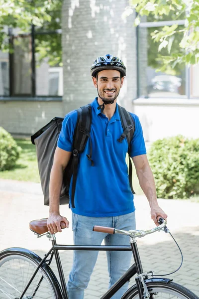 Livreur musulman souriant avec sac à dos marchant près du vélo à l'extérieur — Photo de stock