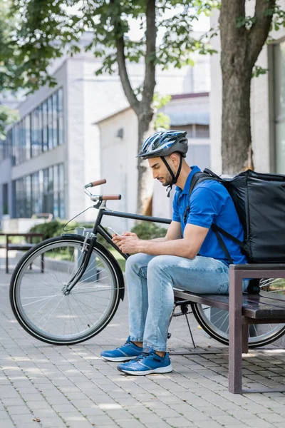 Vista lateral do correio muçulmano em camiseta azul e mochila térmica usando celular no banco perto da bicicleta — Fotografia de Stock