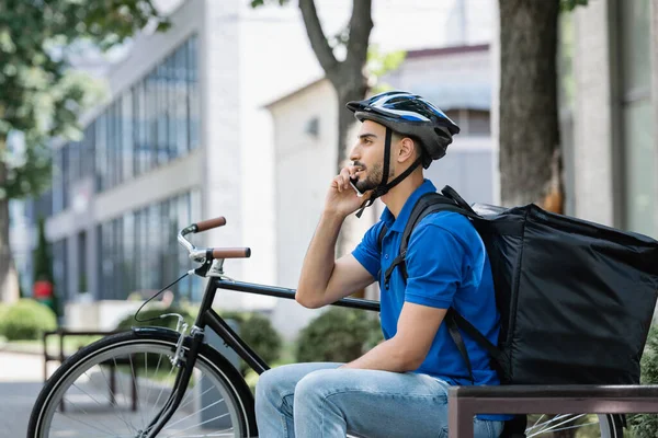 Vista lateral del repartidor musulmán con la mochila termo hablando en el teléfono inteligente cerca de la bicicleta al aire libre — Stock Photo