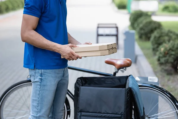 Ausgeschnittene Ansicht von Kurier, der Pizzakartons in der Nähe von geöffnetem Rucksack und verschwommenem Fahrrad hält — Stockfoto