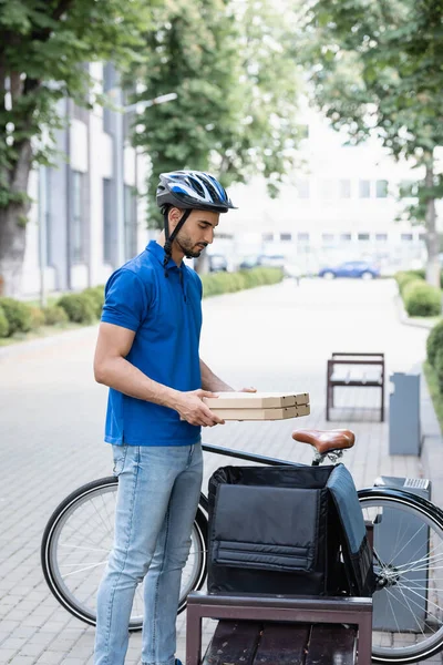 Side view of arabian deliveryman holding pizza boxes near thermo backpack and bike outdoors — Stock Photo