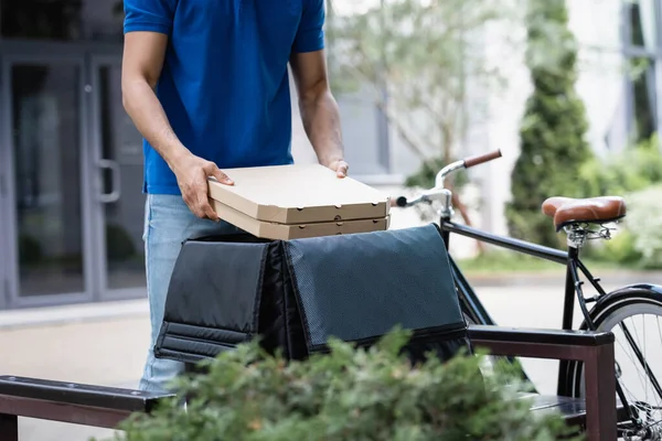 Cropped view of deliveryman taking pizza boxes from thermo backpack on bench and bike outdoors — Stock Photo