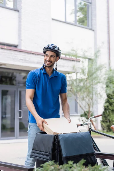 Feliz mensajero árabe en casco tomando cajas de pizza de la mochila termo cerca de la bicicleta en la calle urbana - foto de stock