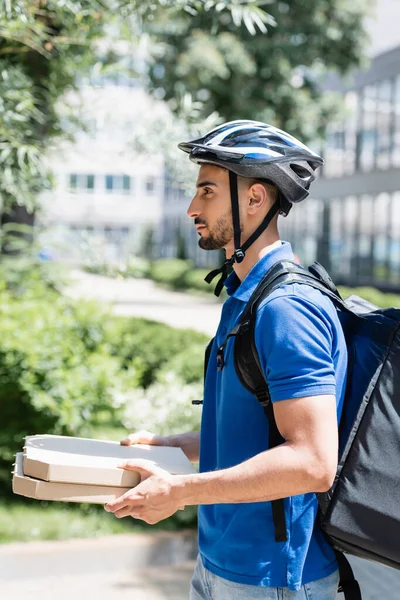 Vista lateral del joven repartidor musulmán en casco sosteniendo cajas de pizza en la calle urbana - foto de stock