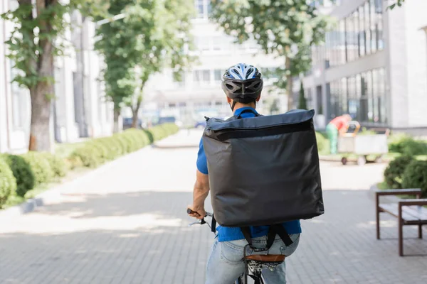 Back view of courier in helmet and thermo backpack riding bicycle on urban street — Stock Photo