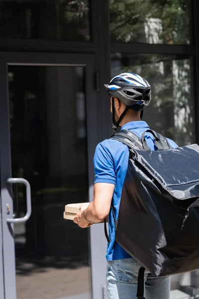 Jovem mensageiro em capacete de segurança segurando caixas de pizza perto da porta do edifício — Fotografia de Stock