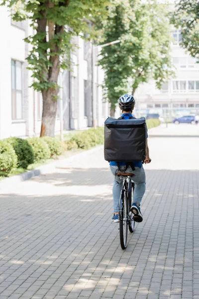 Back view of courier with thermo backpack riding bike on urban street — Stock Photo