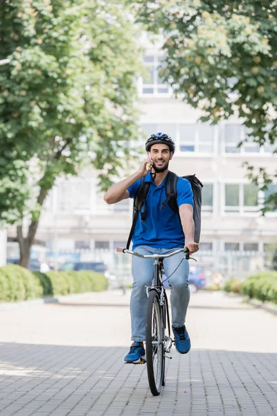 Mensajero musulmán positivo hablando en el teléfono inteligente mientras monta bilis en la calle urbana - foto de stock