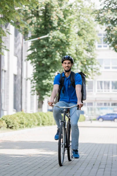 Happy arabian courier in safety helmet and backpack riding bike on urban street — Stock Photo