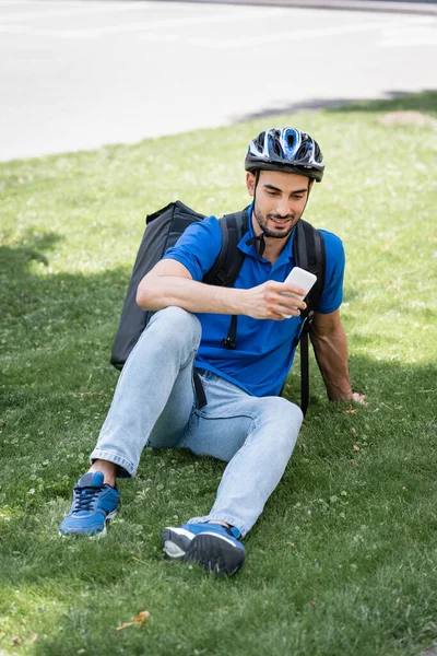 Mensajero musulmán sonriente en casco y mochila térmica usando teléfono inteligente en el césped - foto de stock
