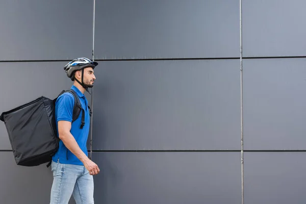 Side view of arabian courier in helmet and thermo backpack walking near building — Stock Photo