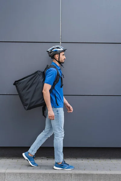 Side view of muslim courier in safety helmet and backpack walking near building — Stock Photo