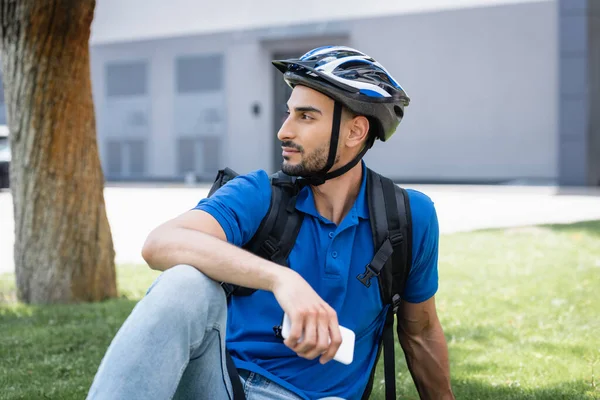 Vue latérale du livreur musulman en casque de protection tenant smartphone sur la pelouse — Photo de stock