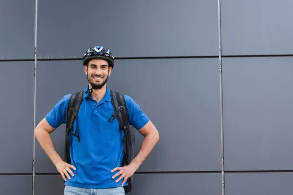 Mensajero árabe sonriente con casco y mochila de pie cerca del edificio al aire libre - foto de stock