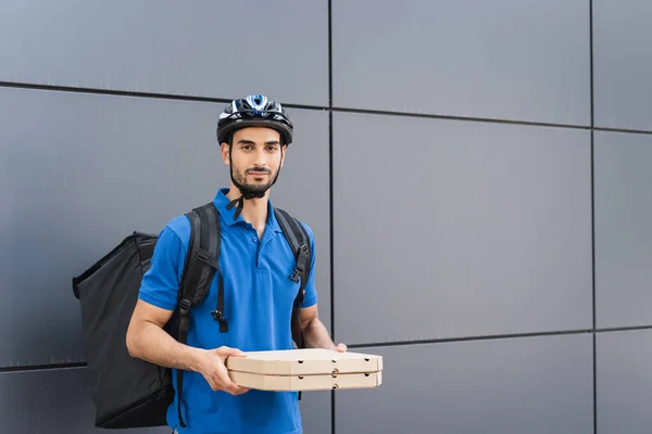 Арабский курьер в шлеме держит коробки с пиццей возле здания на открытом воздухе — стоковое фото