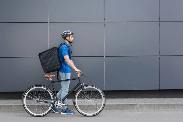 Side view of arabian courier with thermo backpack walking near bike and building outdoors — Stock Photo