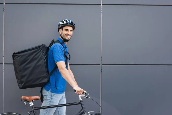 Cheerful muslim deliveryman with backpack and bike walking near building outdoors — Stock Photo