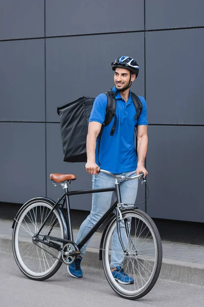 Smiling muslim deliveryman with backpack walking near building on urban street — Stock Photo