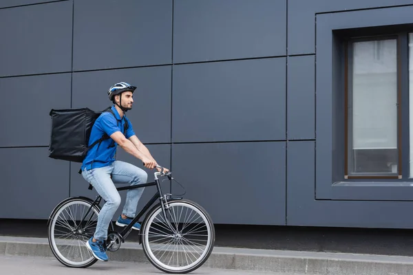 Arabian deliveryman with backpack riding bike on urban street — Stock Photo