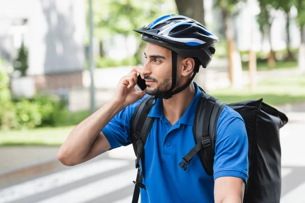 Joven repartidor musulmán en casco de seguridad hablando en el teléfono inteligente al aire libre - foto de stock