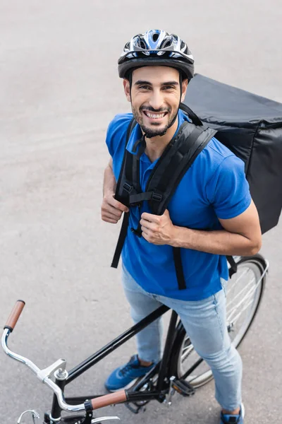 Vista de ángulo alto del alegre repartidor musulmán de pie cerca de la bicicleta al aire libre - foto de stock