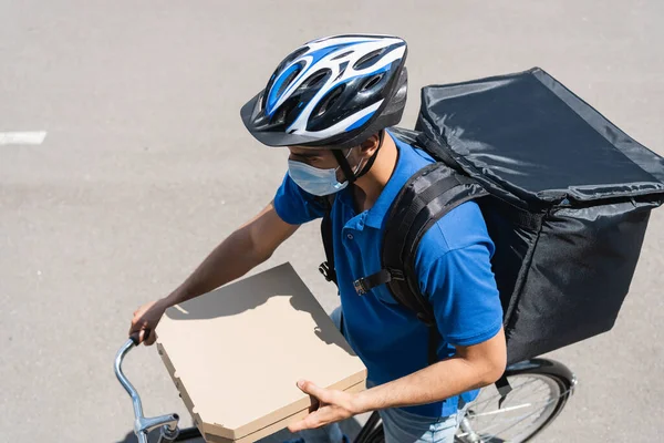 Vista aérea del repartidor musulmán en máscara médica sosteniendo cajas de pizza mientras monta bicicleta - foto de stock