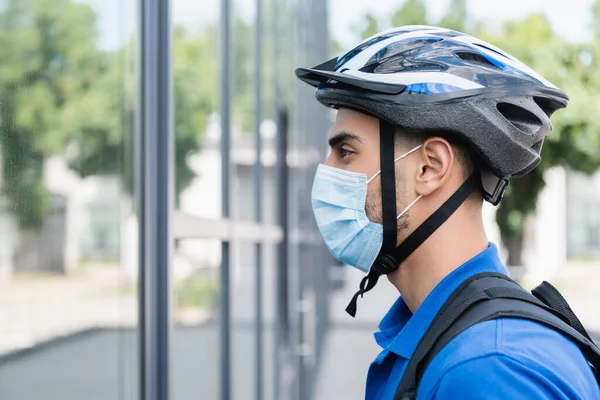 Vista laterale del corriere musulmano in maschera medica e casco da bici in piedi vicino all'edificio sfocato — Foto stock
